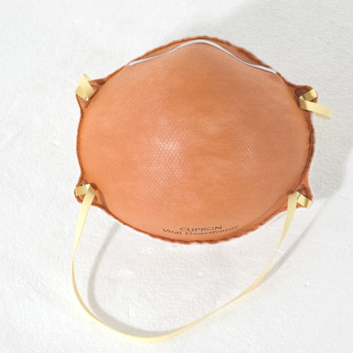 An orange protective face mask with yellow elastic straps lies on a white surface. The mask has text on it that reads Cupron Viral Deactivator.