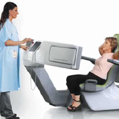 A female patient sitting in a doctors office in a pink shirt and black pants, and a female doctor in a blue lab coat speaking to the patient.