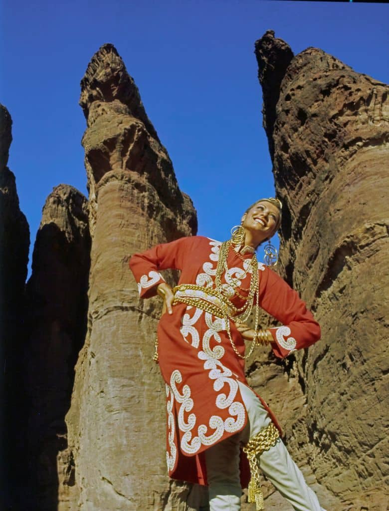 Model Susan Schoenberg poses with her hands on her hips and her legs crossed in front of a large stone outcropping. She is wearing a red caftan in wool-and-rayon with gold braid embellishment by Maskit over tan trousers. Accessories: chains at neck, waist, and knee by Napier, Monet, and Accessocraft; Mark XIV gold bracelet; Richard Palan golden ring earrings. Hair by Carita.  (Photo by John Cowan/Conde Nast via Getty Images)