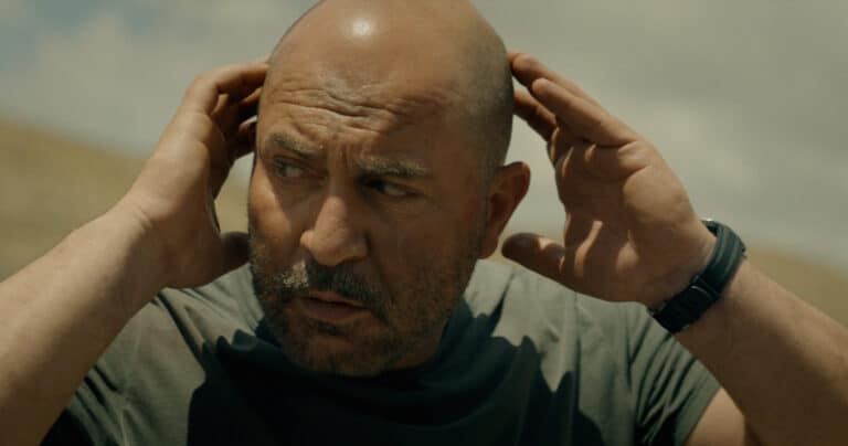 "Bald man with a beard outdoors under a cloudy sky, wearing a dark t-shirt and watch, holding his hands to his head and looking concerned or focused in a natural setting."