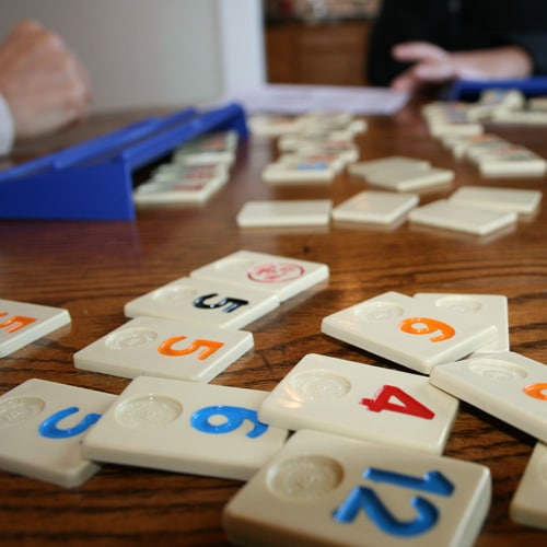 Close-up of a table with a Rummikub game in progress. Tiles with numbers in various colors are scattered, with some placed on blue tile racks. Two people are partially visible, engaged in the game.