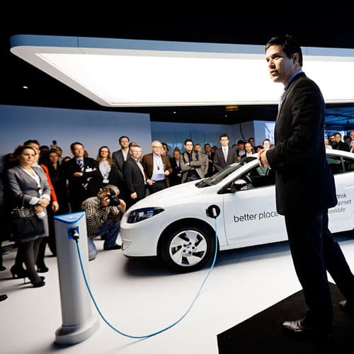A person in a suit stands on a stage beside an electric car with better place on it, plugged into a charging station. A group of people watch in the background. The setting appears to be a modern, indoor presentation.