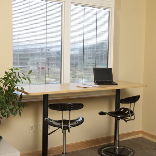 A modern, clean office space with a light wood desk, two black stools, and a laptop. Large windows with blinds let in natural light. A potted plant adds a touch of greenery.